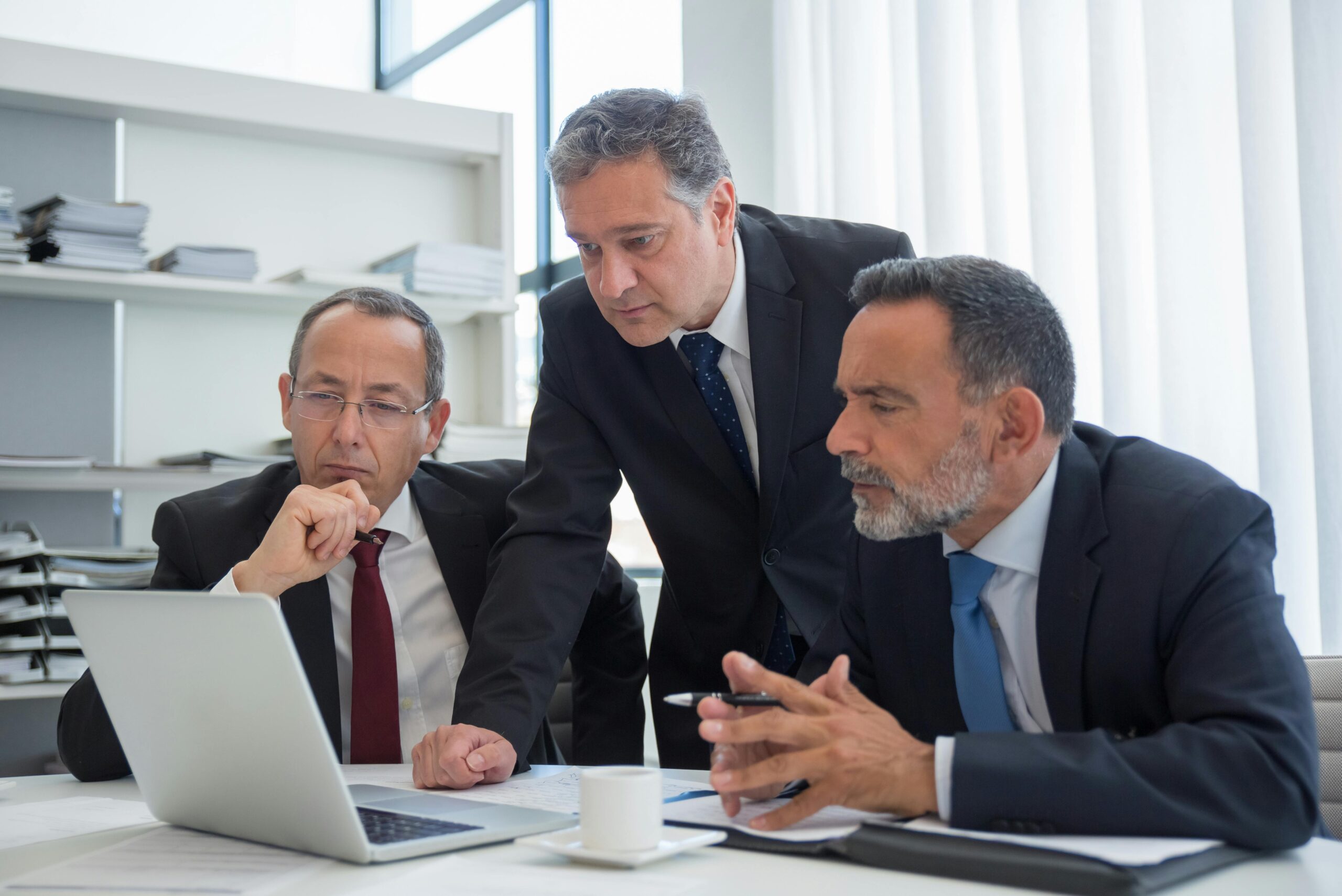 Three businessmen in suits collaborating in a modern office setting, focused on a laptop.